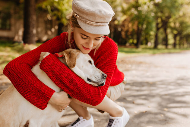 肖像戴着时髦帽子和白色运动鞋的漂亮女孩温柔地拥抱她的狗可爱的金发女郎和她的宠物在公园里玩耍头发公园女人