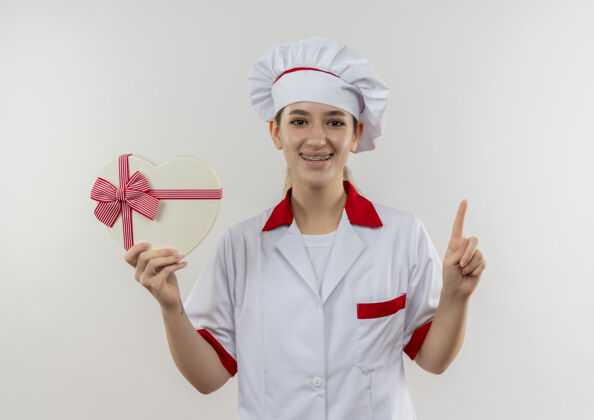 持有微笑的年轻漂亮厨师身着厨师制服 戴着牙套 手持心形礼盒 竖起手指 与白色空间隔离礼品漂亮微笑