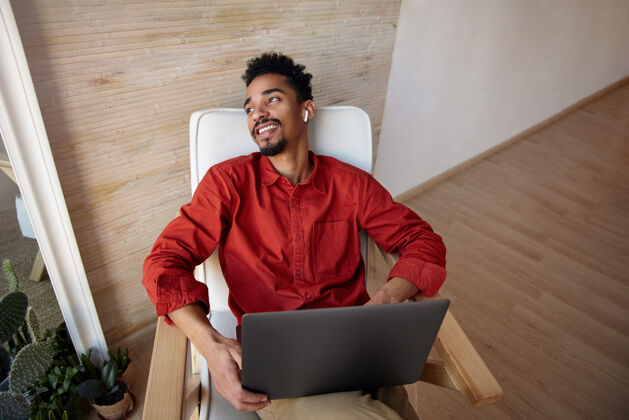工作积极的年轻黑发胡子黑皮肤的家伙在红色衬衫后仰着他的头 而坐在椅子上 愉快地看着窗外 孤立在家里的内部年轻笔记本电脑米色