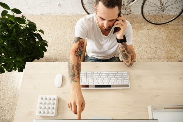 自由职业者纹身男子的顶视图显示了一些东西在显示器上 而在他的工作桌上讲电话在共同工作中心肖像聪明外观