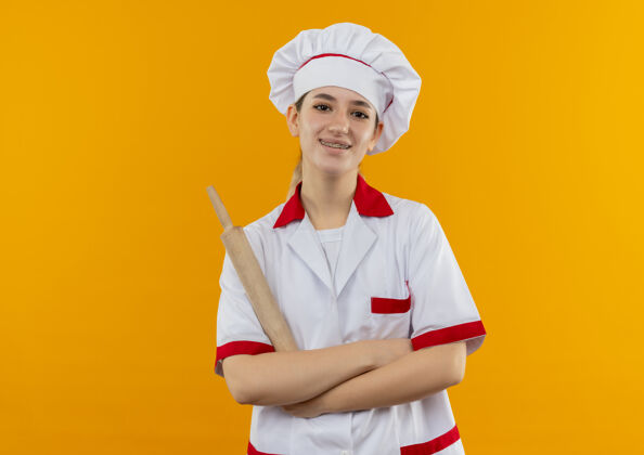 复制微笑着的年轻漂亮厨师身着厨师制服 站着封闭的姿势 拿着擀面杖孤立地站在橙色的空间里厨师别针空间