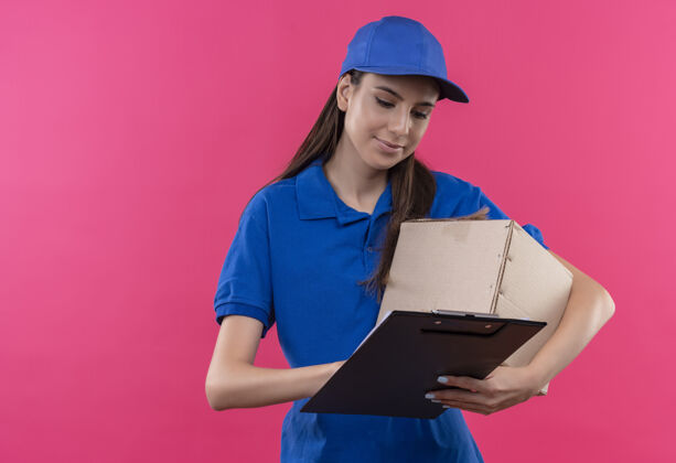 盒子穿着蓝色制服 戴着帽子的年轻送货女孩拿着盒子和剪贴板 严肃地看着空白页剪贴板拿着脸