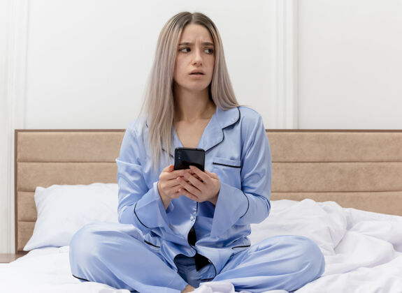坐着穿着蓝色睡衣的年轻美女坐在床上 用智能手机在卧室的灯光背景下困惑地看着旁边智能手机蓝色睡衣