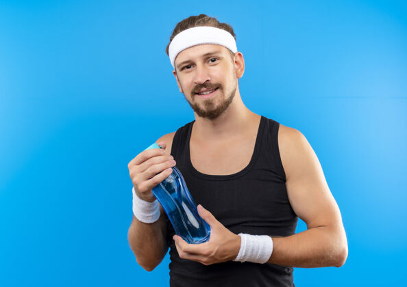 蓝色高兴的年轻英俊的运动型男子戴着头带和手镯拿着水瓶看起来孤立的蓝色空间持有空间高兴