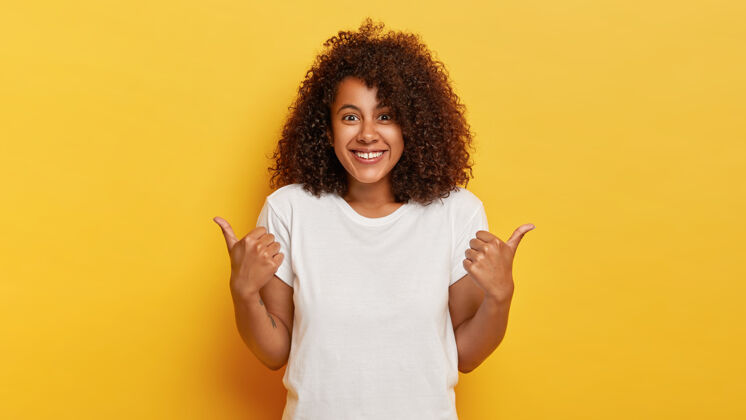 非洲快乐的卷发女孩竖起大拇指 表示对某人的支持和尊重 愉快地微笑 达到理想的目标 穿着白色t恤 隔离在黄色的墙上微笑欢笑特写