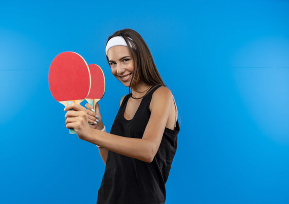 乒乓球带着微笑的年轻漂亮的运动女孩戴着头带和腕带拿着乒乓球拍孤立在蓝色的空间蓝色女孩持有