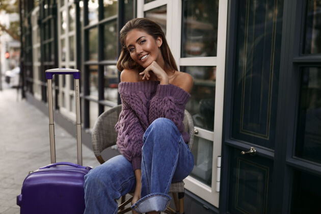 紫色漂亮女孩拿着手提箱坐在咖啡馆附近的椅子上 穿着紫色套头衫 牛仔裤 化妆 发型 情感 秋天 金发 快乐 针织 微笑 调情休息街道头发