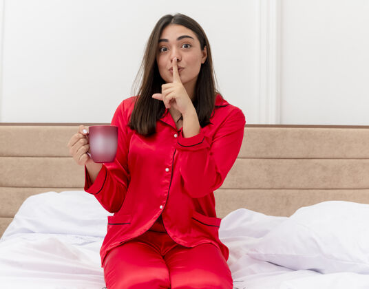 沉默穿着红色睡衣的年轻漂亮女人坐在床上喝着咖啡 手指放在嘴唇上做着沉默的手势 卧室内部的灯光背景下睡衣手指室内
