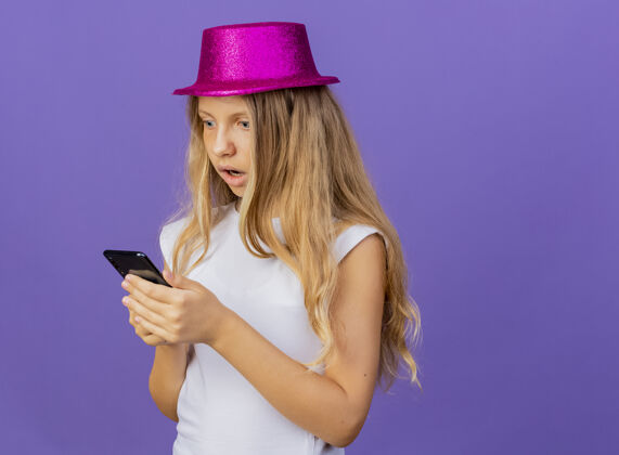 女孩戴着节日礼帽的漂亮小女孩用智能手机惊诧不已 生日派对概念站在紫色背景上生日节日惊人