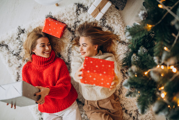 拿着礼物两个女孩朋友坐在树边拿着圣诞礼物传统乐趣节日