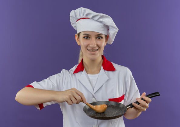 制服微笑着的年轻漂亮的厨师 穿着厨师制服 戴着牙套 把煎锅和勺子隔在紫色的空间里年轻牙科薯条