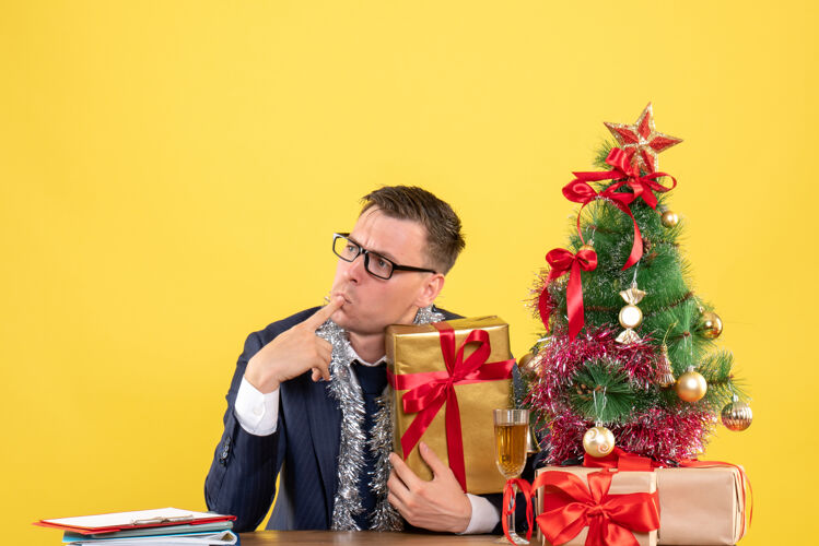 新郎体贴的男人把手指放在嘴上坐在圣诞树旁的桌子前 黄色的礼物桌子体贴男人