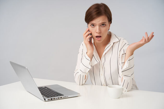 女士困惑的年轻棕色眼睛短发黑发女子与自然化妆有着愉快的电话交谈 而坐在桌边的现代笔记本电脑衣服工作会议