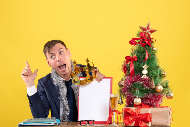 生意人商务人士拿着剪贴板和皇冠坐在圣诞树旁的桌子前 黄色的礼物黄色礼物圣诞节
