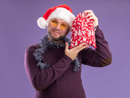 穿着高兴的中年男子戴着圣诞帽 脖子上戴着金箔花环 戴着眼镜 头上拿着圣诞礼品袋 孤零零地站在紫色的墙上头高兴周围