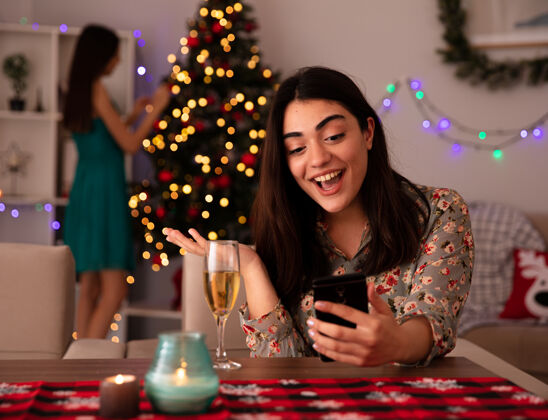 圣诞快乐高兴的年轻女孩装饰圣诞树 她惊讶的朋友看着坐在桌边的手机 享受着家里的圣诞节时光快乐看电话