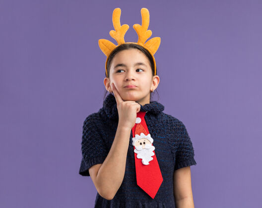 紫穿着针织连衣裙的小女孩戴着红领带 边上有滑稽的镶边 头上有鹿角 困惑地看着一边年新新年