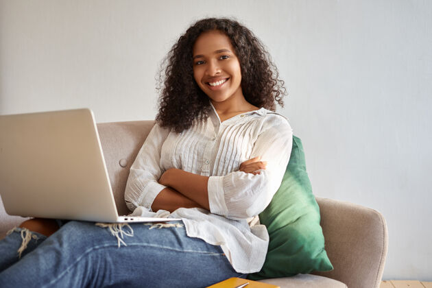 年轻人美丽开朗自信的非洲裔美国年轻女子坐在沙发上 手里拿着笔记本电脑 双臂交叉放在胸前 看着网上的视频博客 笑容满面冲浪非洲使用