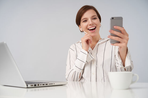 耳机快乐的年轻短发黑发女性 随意的发型 举着手机打视频电话 坐在白色的椅子上开心地笑着化妆肖像工作