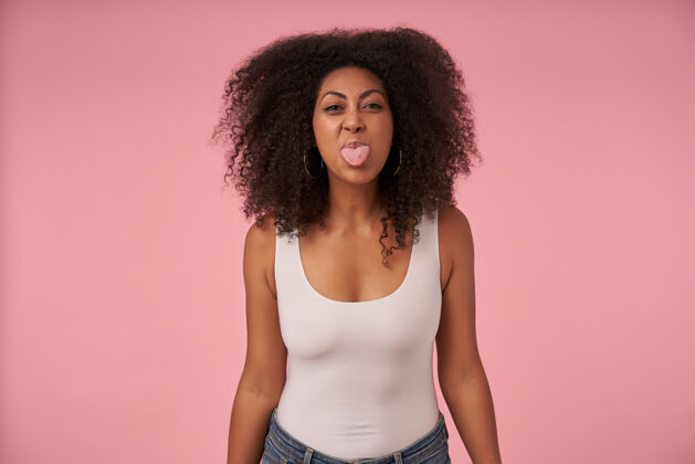 非洲年轻的黑皮肤女性的肖像 随意的发型 穿着白衬衫和牛仔裤 愉快地露出舌头 孤立的粉红色黑色化妆语言
