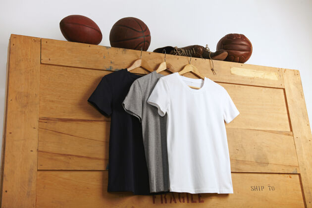 牛仔裤展示一件白色 灰色和黑色的普通短袖t恤 在木制的运输箱上放着复古的足球 篮球和排球以及旧的运动皮靴时尚健身房休闲