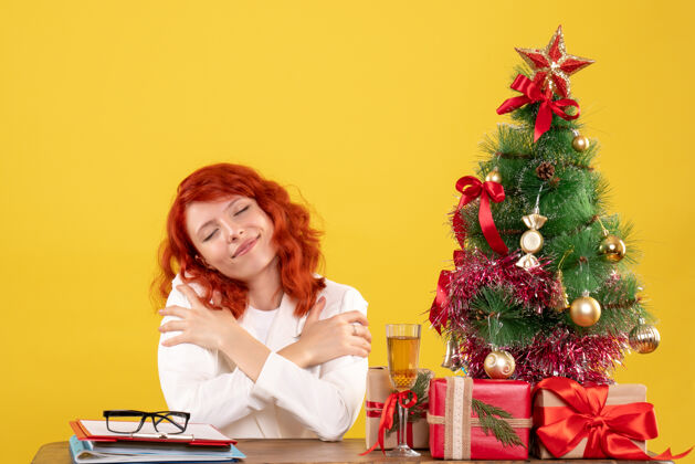 女性前视图：女医生坐在桌子后面 黄色背景上有圣诞礼物 圣诞树和礼品盒桌子医生情感