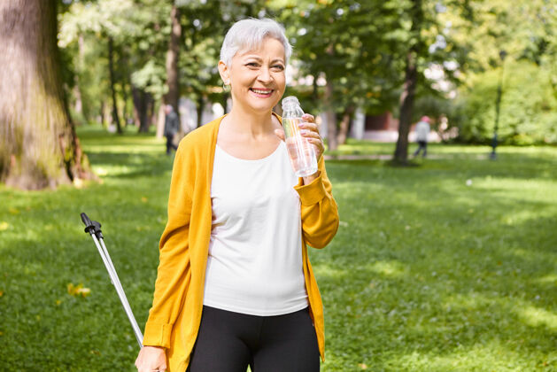 运动美丽健康的老年妇女肖像 长着一头精灵般的灰色头发 在公园里散步时休息 用北欧的斯堪的纳维亚的电线杆 拿着瓶子 喝着水 感觉精力充沛 面带微笑健身老年人北欧