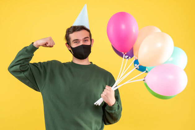 气球正面图：戴着派对帽的年轻人 五颜六色的气球展示着站在黄色地面上的肌肉男人生日年轻