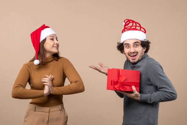 帽子新年气氛喜庆的概念与有趣可爱的情侣戴着红色圣诞老人帽女孩送他的礼物背后的灰色帽子圣诞节礼物