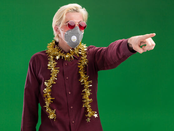 圣诞节年轻的金发男子戴着眼镜 戴着防护面具 脖子上戴着金属丝花环 看着并指着隔离在绿色墙上的一面金发男人年轻