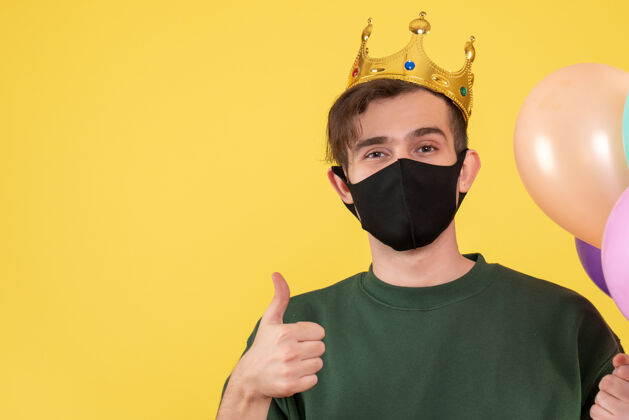 防护正面图：戴着皇冠和黑色面具的年轻人在黄色上做拇指朝上的标志皇冠伪装年轻人