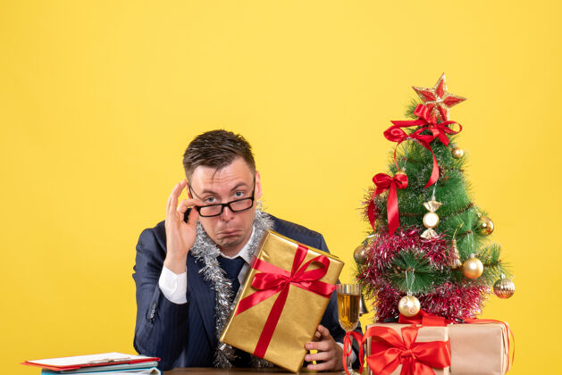 圣诞节戴眼镜的帅哥坐在圣诞树旁的桌子前 黄色的礼物新郎商人男