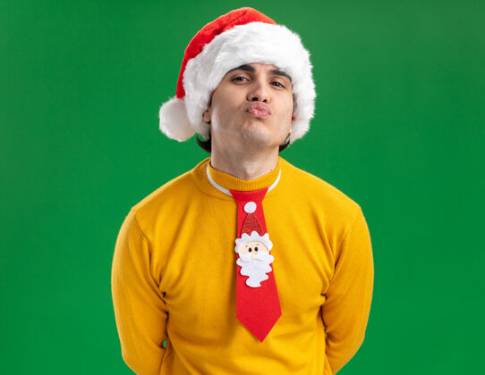 男人快乐的年轻人穿着黄色高领毛衣 戴着圣诞老人帽 打着有趣的领带 站在绿色的背景上 嘴唇就像要亲吻一样年轻亲吻嘴唇