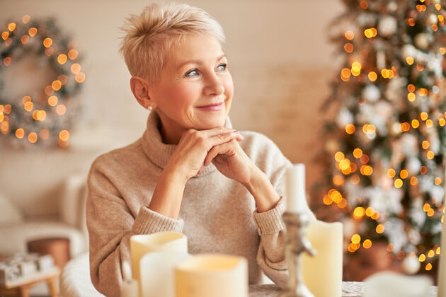 季节穿着大号毛衣的安详时尚的中年白人妇女 有着沉思梦幻的表情 微笑着 拿着蜡烛坐在桌子旁 等待朋友们来庆祝平安夜夜晚圣诞节华丽