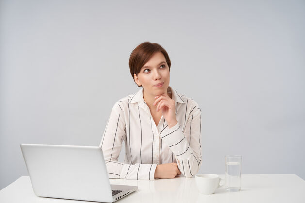 玻璃沉思的年轻可爱的棕色头发的女性 随意的发型积极地看着一边 下巴靠在举起的手上 穿着正式的衣服 在白色的脸上摆姿势坐着笔记本电脑条纹