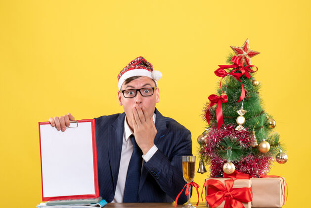 男人举着夹子坐在圣诞树旁的桌子旁 拿着黄色礼物的大眼睛商人的正面图桌子商人礼物