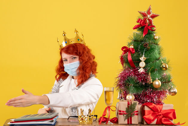 穿正面图：女医生拿着圣诞礼物 戴着皇冠坐在黄色的桌子上花快乐礼物