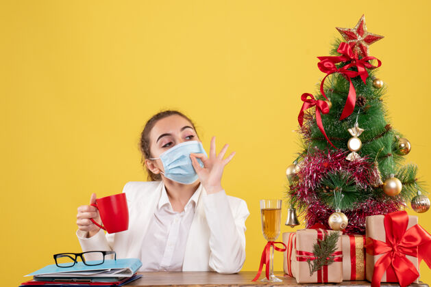 前面正面图女医生戴着防护面具坐在黄色背景上 戴着圣诞树和礼品盒人外套实验室