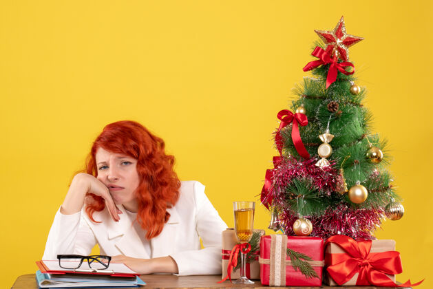 无聊前视图：女医生坐在桌子后面 黄色背景上有圣诞树和礼品盒 里面有圣诞礼物人快乐年