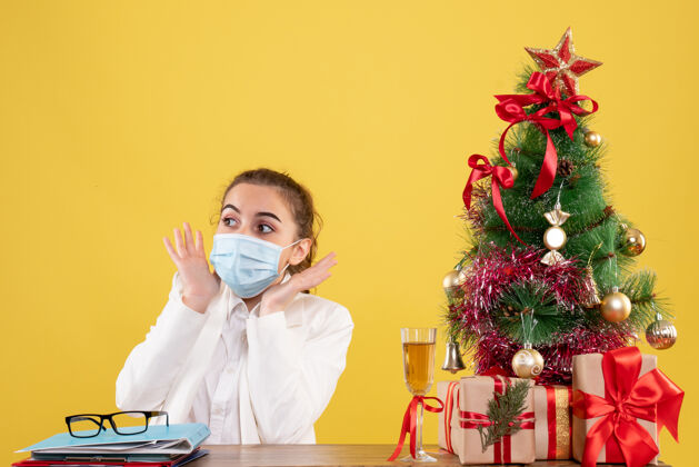 医生前视图女医生戴着防护面具坐在黄色背景上 带着圣诞树和礼品盒保护背景坐着