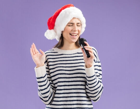 女孩戴着圣诞帽的年轻白人女孩拿着麦克风 假装在紫色背景上唱着与世隔绝的歌曲唱新的新年