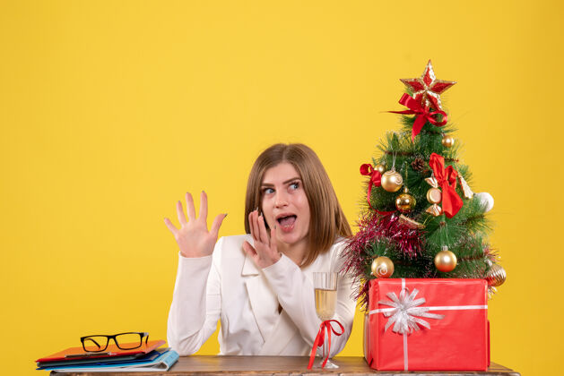 快乐前视图：女医生坐在桌子前 拿着圣诞礼物 黄色背景上的树医生情感背景