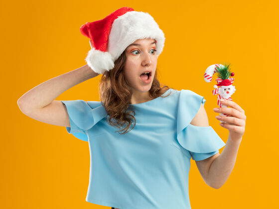 顶身穿蓝色上衣 戴圣诞帽的年轻女子手持圣诞糖果手杖 手放在头上惊奇地看着它年轻帽子手杖