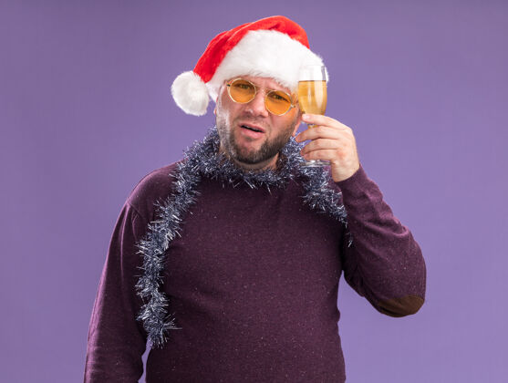 穿着中年男子戴着圣诞帽 脖子上戴着金箔花环 戴着一副眼镜 手里拿着一杯香槟 隔离在紫色的墙上圣诞节脖子帽子