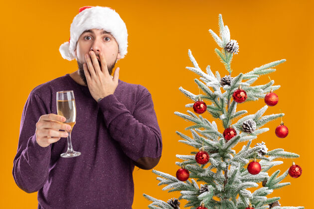 帽子一个穿着紫色毛衣 戴着圣诞帽 手里拿着一杯香槟的年轻人站在橙色墙上的圣诞树旁 惊讶不已玻璃杯站着圣诞老人