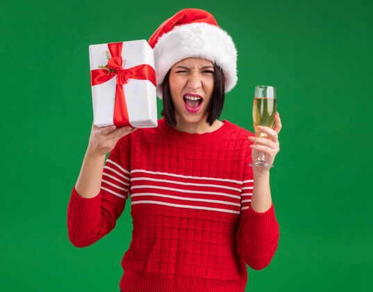 圣诞老人快乐的年轻女孩戴着圣诞帽拿着礼包靠近头部和一杯香槟看着相机眨眼孤立的绿色背景年轻穿着包装