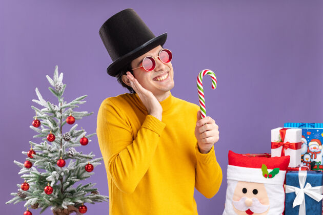高领毛衣快乐的年轻人穿着黄色高领毛衣 戴着黑色帽子 手持糖果手杖 站在一棵圣诞树旁 愉快地微笑着 紫色背景下的礼物礼物拿着男人