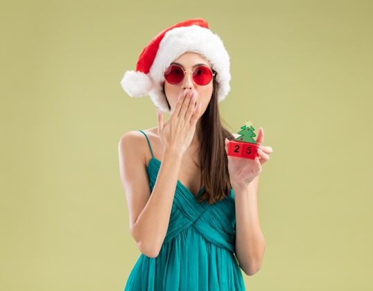 新一个戴着太阳眼镜 戴着圣诞帽的白人女孩 手放在嘴上 手里拿着圣诞树饰物年复制年轻