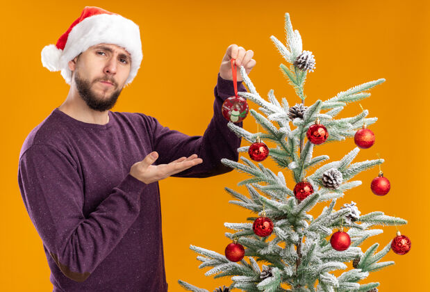 抱一个穿着紫色毛衣 戴着圣诞帽的年轻人站在圣诞树旁 手里拿着一个玩具 把它挂在树上 脸上带着严肃的表情 背景是橙色的挂年轻人认真