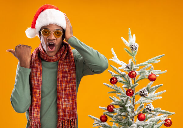 非洲人戴着圣诞帽 脖子上围着围巾的非洲裔美国人看着摄像机 惊讶地用拇指指着旁边站在橙色背景下的圣诞树看脖子惊奇